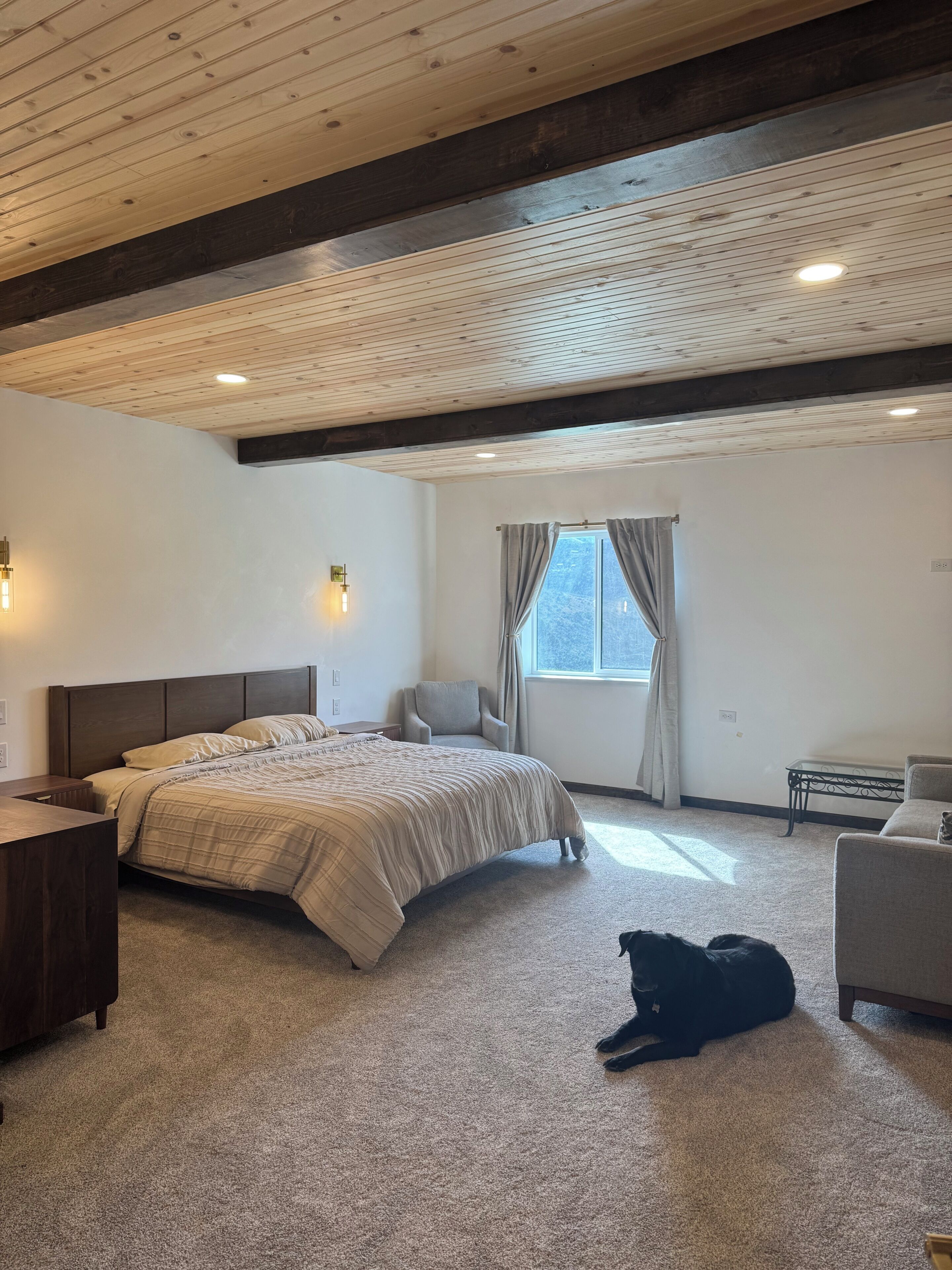 A bedroom with a large bed, a chair, a small table, and a dog lying on the floor.