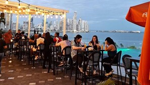 Un área de comedor al aire libre con mesas y sillas, un horizonte urbano al fondo y vista al agua.