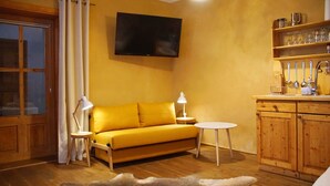 Un salon chaleureux avec un canapé jaune, deux tables d’appoint avec des lampes, un téléviseur à écran plat fixé au mur, des meubles en bois et une petite table ronde.