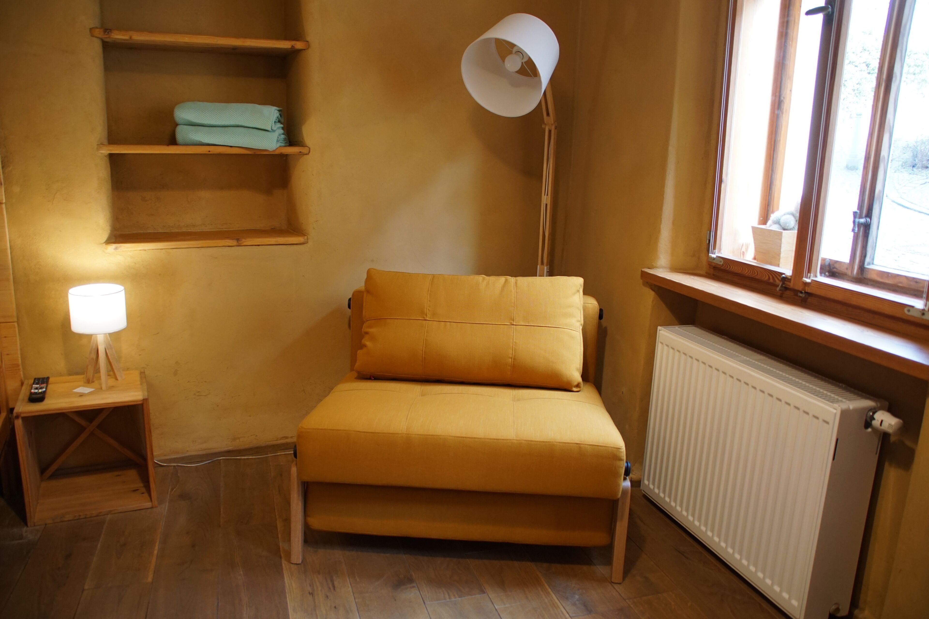 Um quarto pequeno e acolhedor, com uma cama de solteiro, mesa de cabeceira com luminária, prateleira com toalhas dobradas e janela com vista.