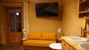 Une pièce chaleureuse avec un plancher en bois, un canapé jaune, une petite table blanche, un téléviseur fixé au mur et un meuble en bois avec un évier.