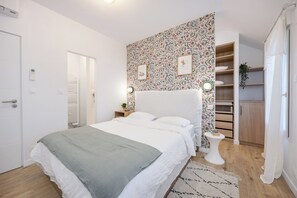 Une chambre à coucher comprenant un lit, des tables de chevet, une commode, un mur recouvert d’un papier peint à motifs, des toilettes et une étagère avec une plante.