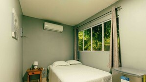 A bedroom with a bed, a nightstand, an air conditioner, and a window with curtains. - Hotel Castillo Blanco (El Zonte)