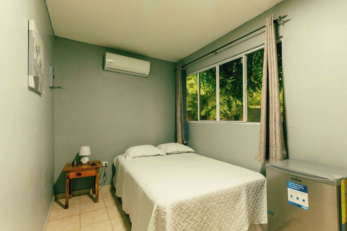 Ein Schlafzimmer mit Bett, Nachttisch, Klimaanlage und einem fenster mit Vorhängen.