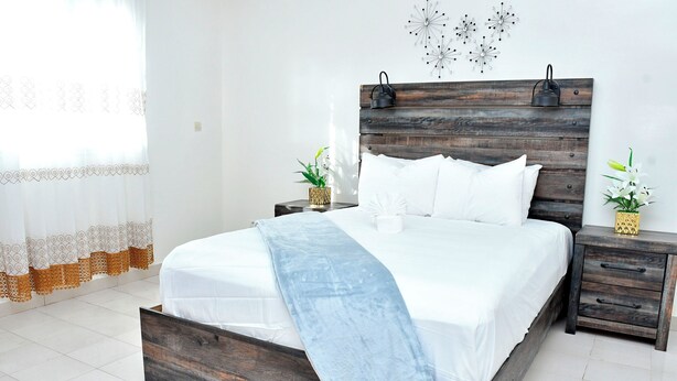 A bedroom with a wooden bed frame, white bedding, a nightstand with a lamp, and decorative wall art. - MAGNIFICENT FURNISHED VILLA, TEMPORARY RENTAL, CITE ALiou Sow Golf Sud (Dakar)