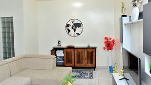 A living room with a sofa, a wooden cabinet, a world map clock, a vase with red flowers, and a flat-screen TV. - MAGNIFICENT FURNISHED VILLA, TEMPORARY RENTAL, CITE ALiou Sow Golf Sud (Dakar)