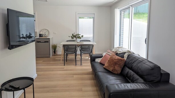 A modern living room with a black leather sofa, a dining table with chairs, a kitchen area, and a TV mounted on the wall.