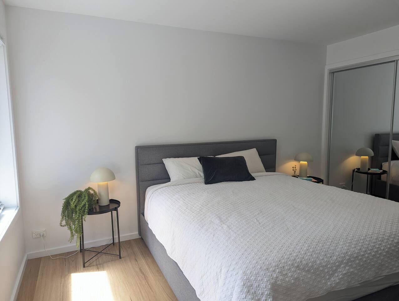 Ein modernes Schlafzimmer mit einem großen Bett, zwei Nachttischen mit Lampen, einer Topfpflanze und einem großen Spiegel an der Wand.