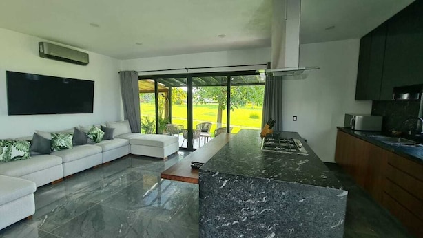 A modern living room with a large sectional sofa, a kitchen area with dark countertops, and a large window offering a view of a green landscape. - Residence near the beach (Mezcales)