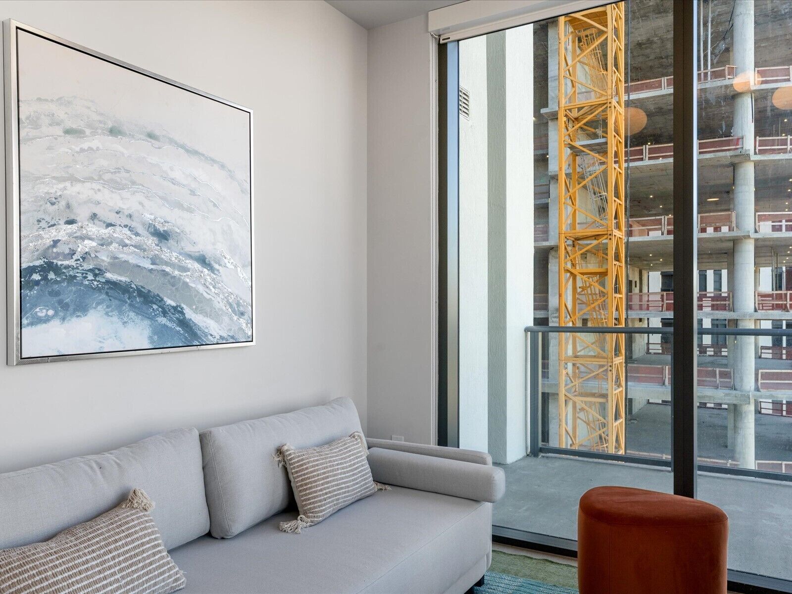 A modern living room with a grey sofa, a large abstract painting, and a view of a construction site through a large window.