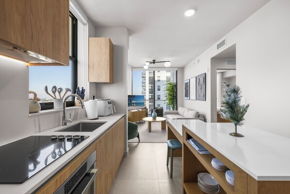 A modern kitchen with a built-in stove, a sink, and wooden cabinets. There is a dining area with a table and chairs, and a view of a building outside. - Downtown Brickell - Desirable Location - We Host (Miami)