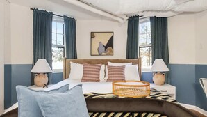 Une chambre à coucher comprenant un lit, deux lampes de chevet, une tête de lit, un tableau abstrait accroché au mur et une fenêtre avec des rideaux.