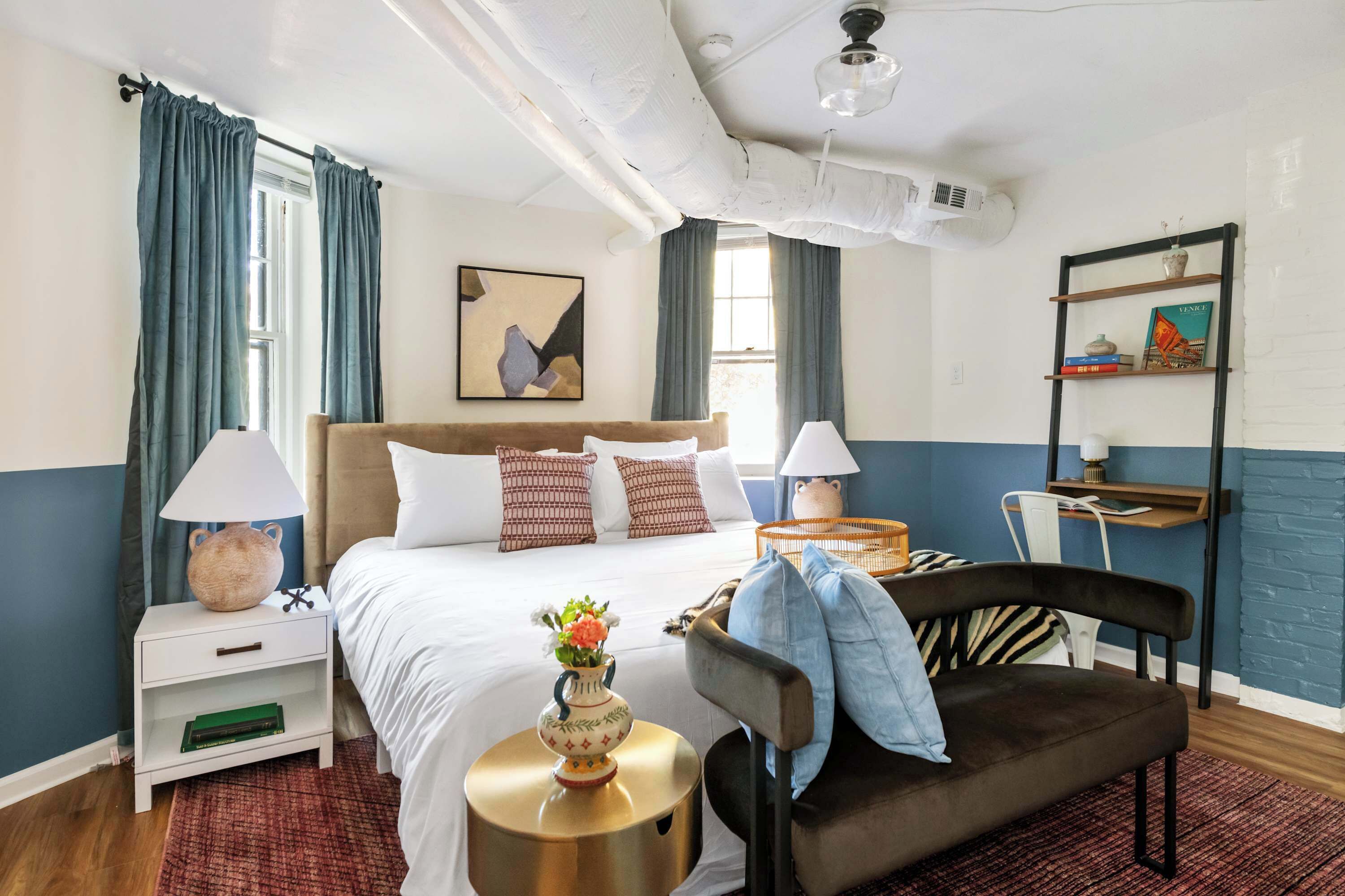 A hotel room with a bed, bedside tables, a chair, a shelf with books, and a painting on the wall.