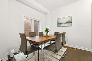 A dining area with a wooden table, six chairs, a potted plant, and a painting on the wall.