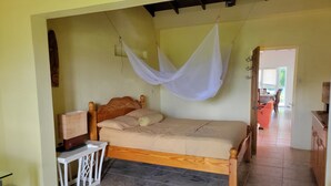 Une chambre avec un lit en bois, une table de chevet, une moustiquaire suspendue et une vue sur la salle à manger adjacente.