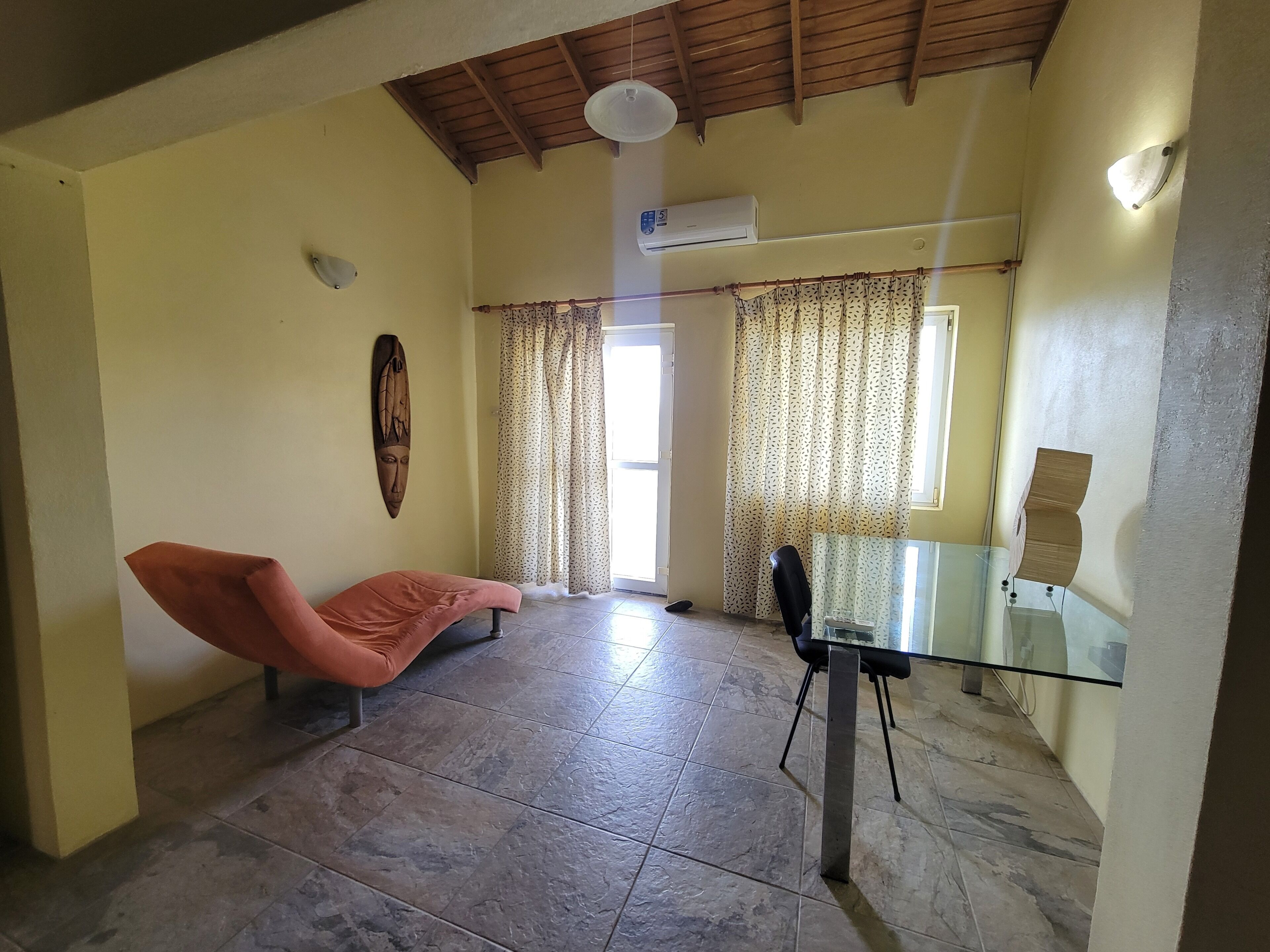 A room with a glass table, a cushioned chair, a ceiling fan, an air conditioning unit, patterned curtains, and a wooden ceiling.