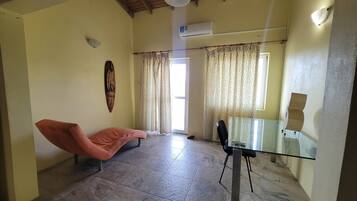 A room with a glass table, a cushioned chair, a ceiling fan, an air conditioning unit, patterned curtains, and a wooden ceiling.