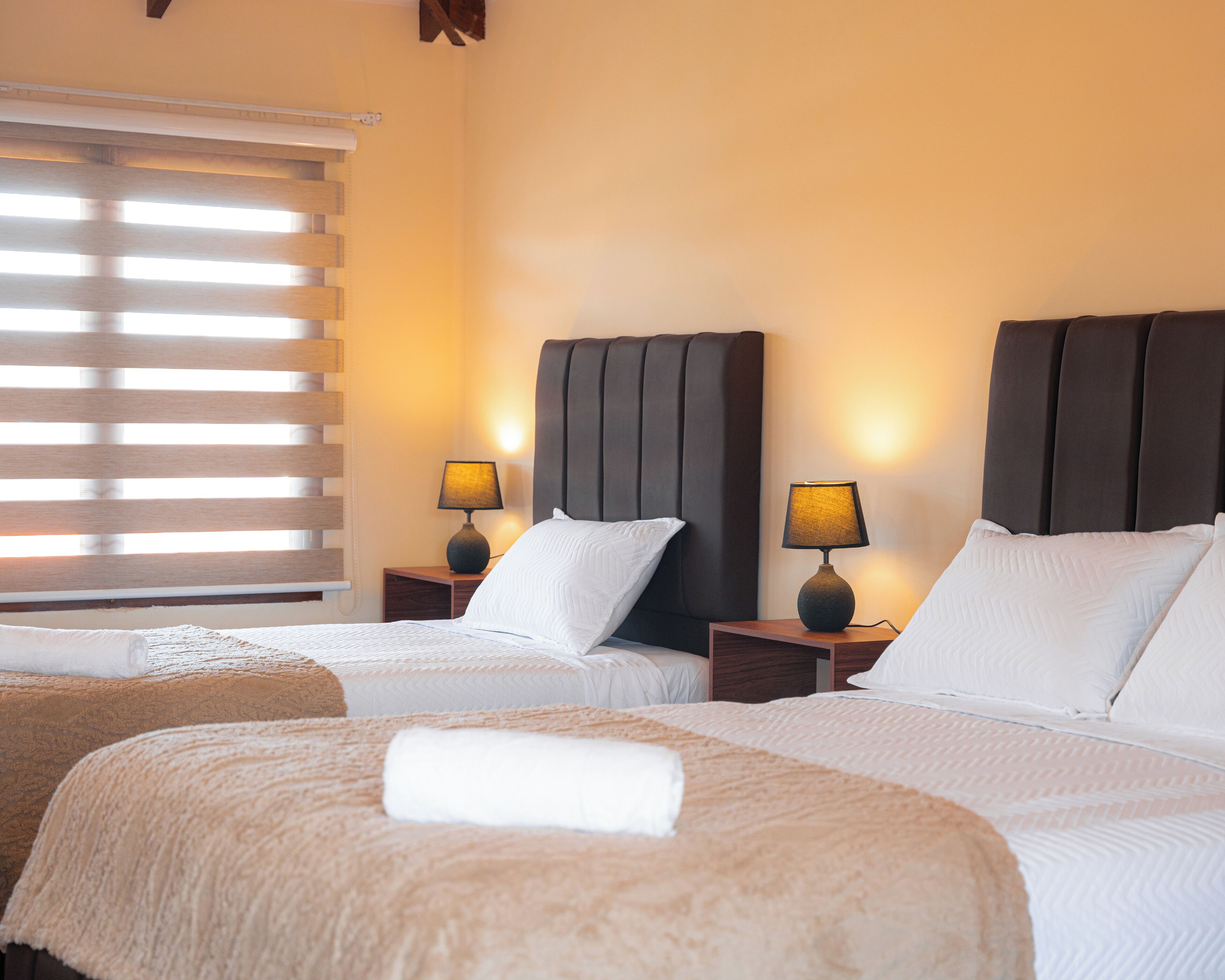 Ein Hotelzimmer mit zwei Betten, jeweils einem Nachttisch und einer Lampe, sowie einem Fenster mit Jalousien.