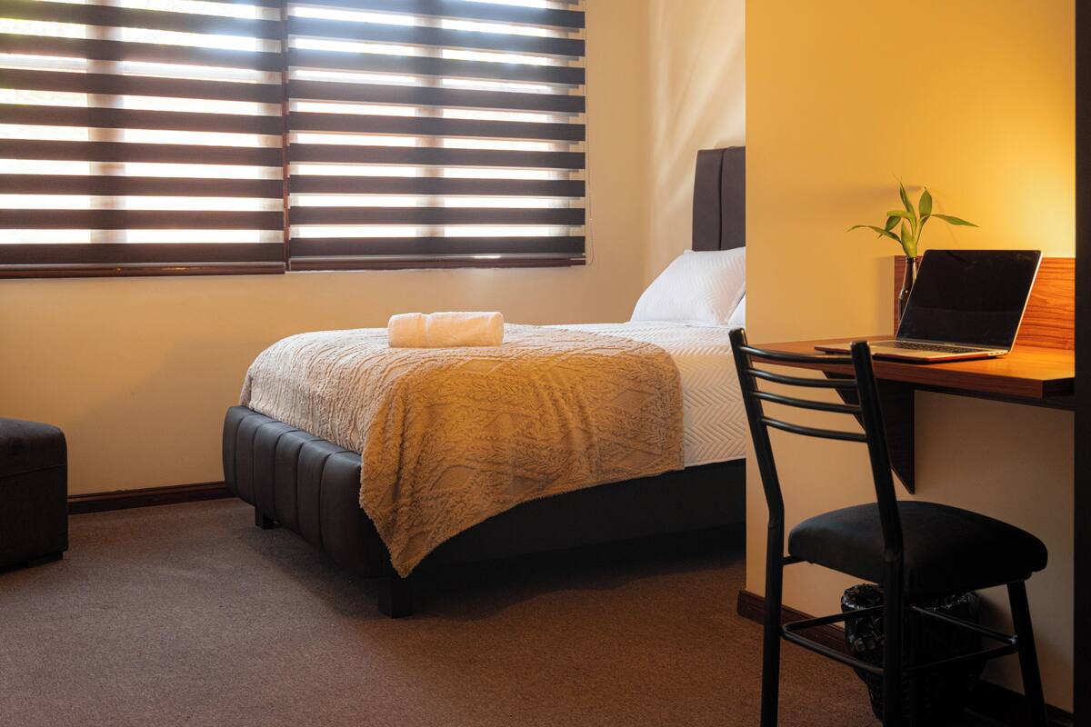 A bedroom with a bed, a desk with a laptop, a chair, a plant, and a window with blinds.