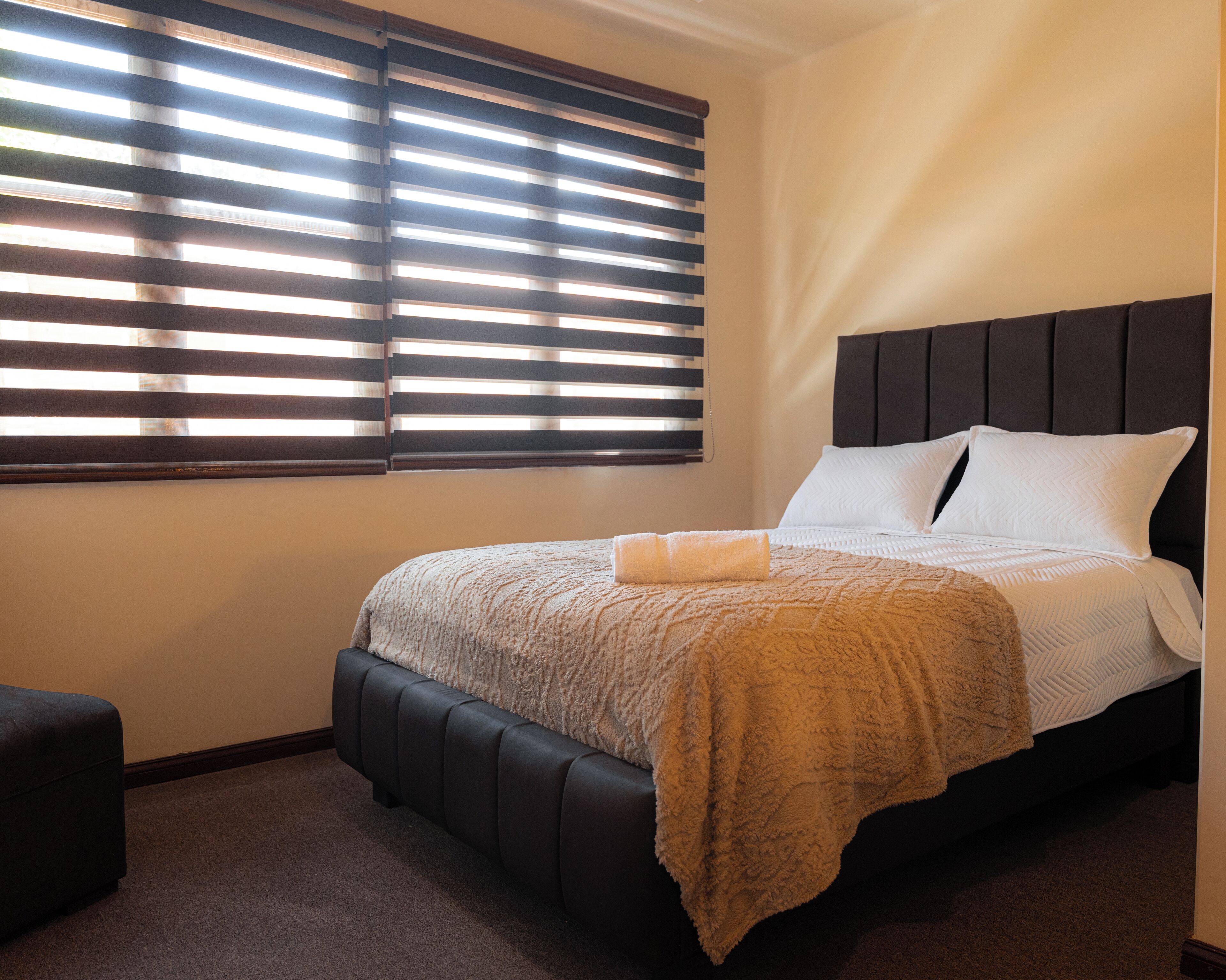 A neatly made bed with a dark headboard, a window with horizontal blinds, and a dark-colored ottoman to the left.