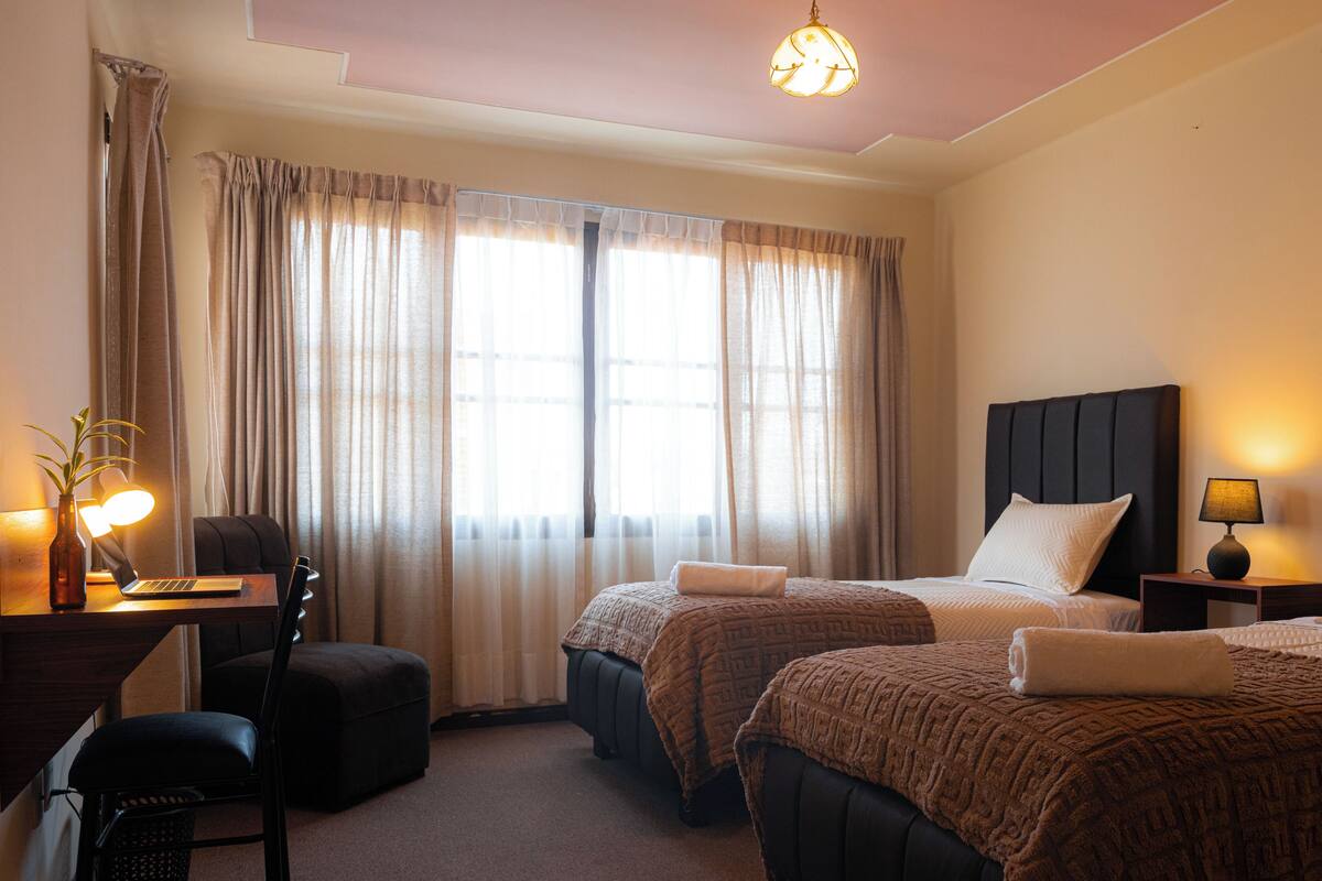 A hotel room with two beds, a desk with a laptop, a chair, a lamp, and a window with curtains.