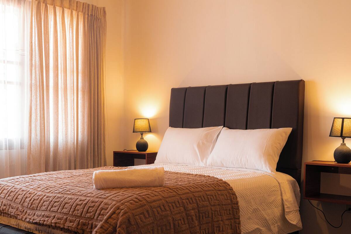 A hotel room with a bed, two bedside tables with lamps, a window with curtains, and a headboard.