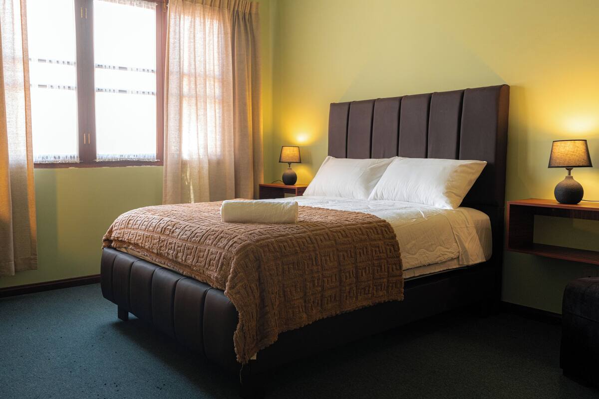 A neatly made bed with a headboard, two bedside lamps, a window with curtains, and a small bedside table.
