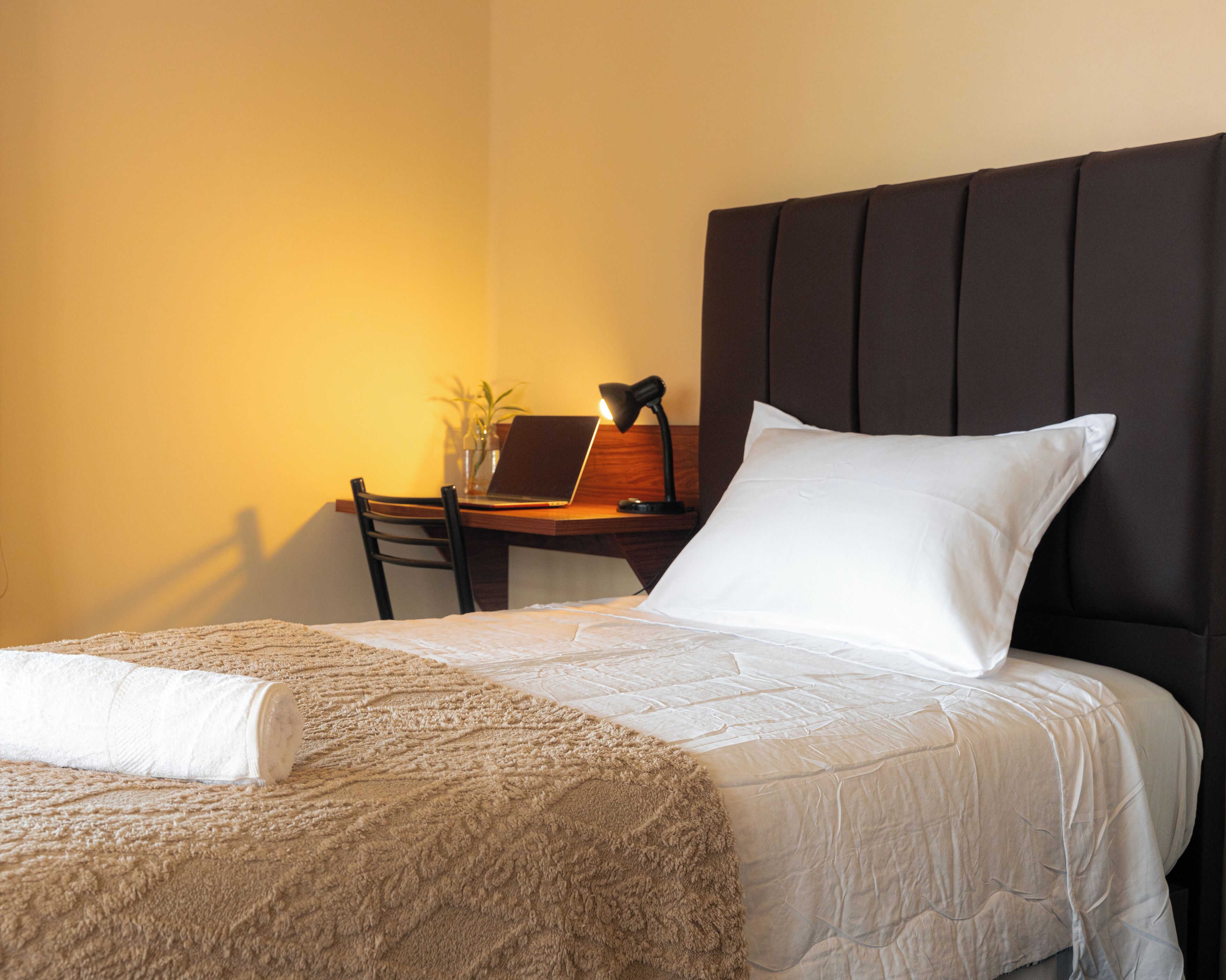 Una cama bien tendida con un almohadón blanco y una manta color beige, un escritorio de madera con una computadora portátil y una lámpara, y una silla.