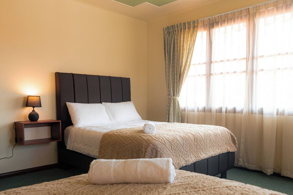 A hotel room with a bed, a bedside table with a lamp, and a window with curtains.