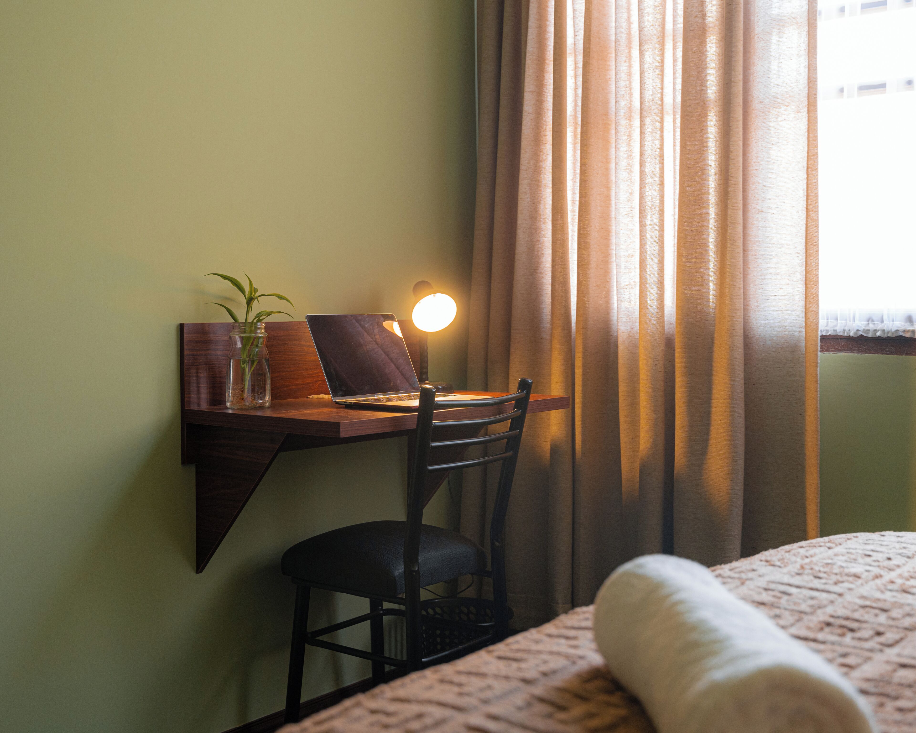 A room with a desk, chair, lamp, and a bed with a blanket.