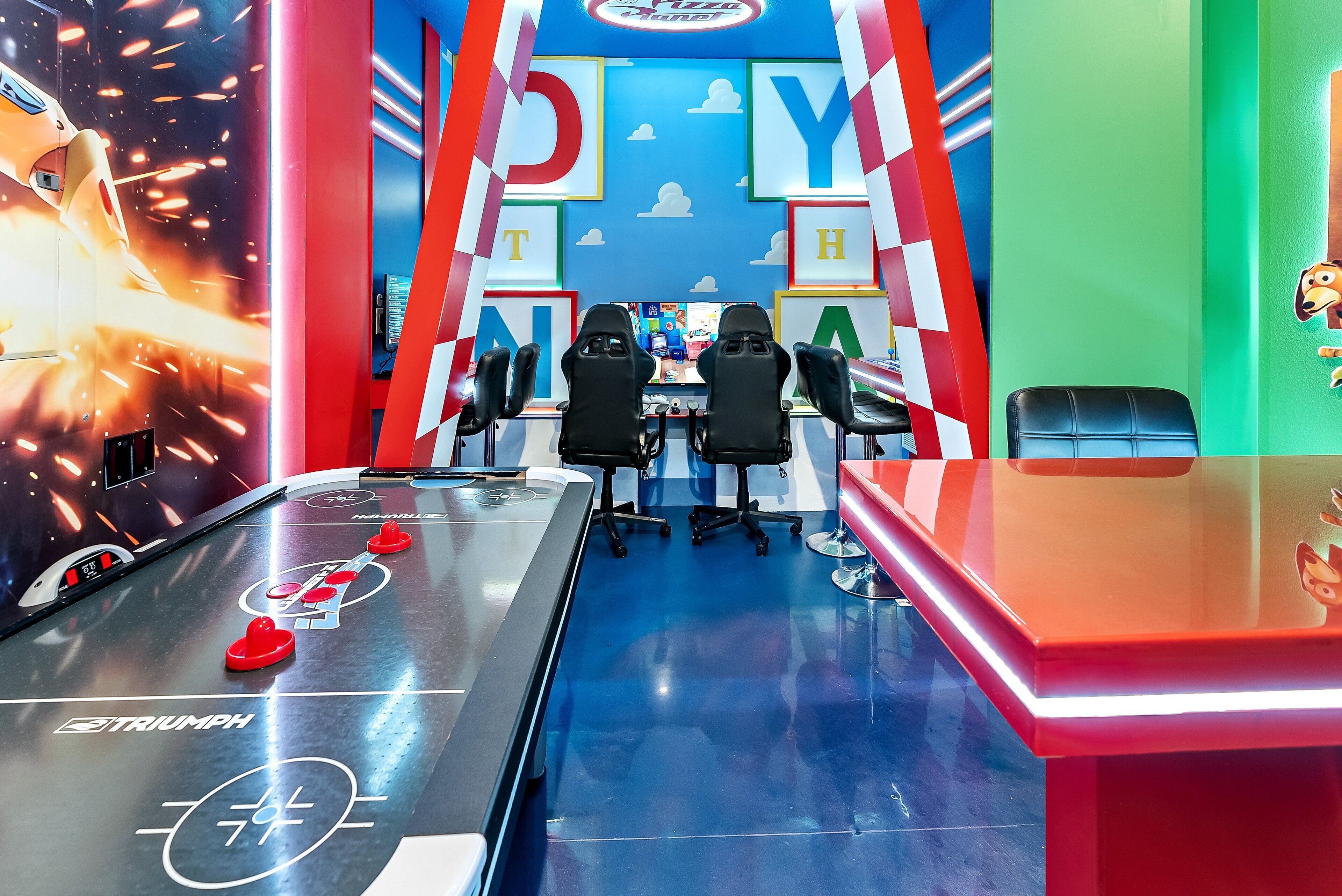 A shuffleboard table with chairs and a neon sign in a vibrant room with colorful walls and a ceiling mural.