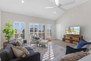 A spacious living room with a ceiling fan, large windows, a sofa, a chair, and a TV on a wooden stand. - Beach access + 3Bedroom + Cozy Patio (Gulf Shores)
