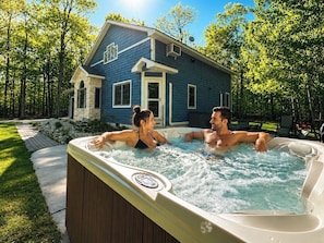 A hot tub with two people in it, a house in the background, and a grassy area with trees. - Private Wooded Retreat w/ Hot Tub • Dog Friendly (Egg Harbor)