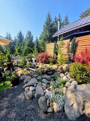 A landscaped garden with a variety of plants and rocks, a wooden building with a sloped roof, and a clear blue sky.