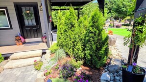 Une maison moderne dotée d’un porche abrité, d’un chemin en pierre et d’un jardin où poussent diverses plantes et fleurs.