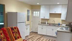 Een knus woongedeelte met een kitchenette, een grijze bank met een kleurrijke deken, en een houten vloer.