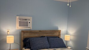 Een slaapkamer met een houten hoofdbord, een gebogen beddeken, twee bedlampen, een plafondventilator en een airconditioner aan de muur.