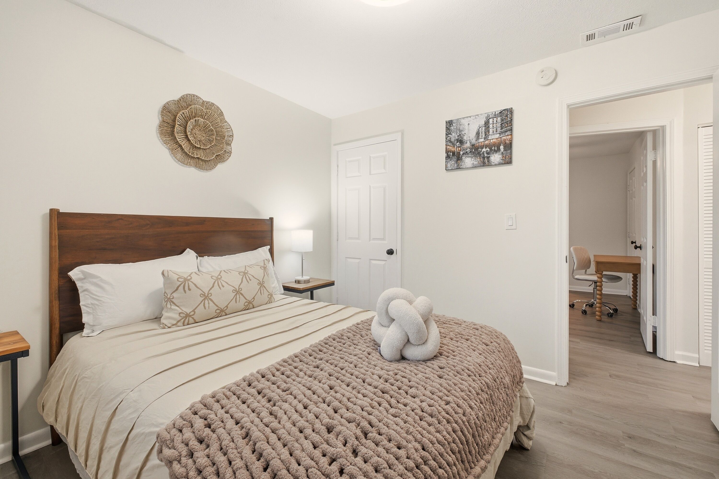 Ein Schlafzimmer mit Holz-Kopfteil, einem großen Bett mit beiger Decke, einem Wandbild und einer Tür zu einem anderen Raum.