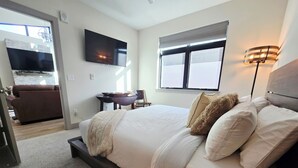 A bedroom with a large bed, a flat-screen TV mounted on the wall, a window with blinds, and a bedside lamp. - Silverthorne Ski Loft - Garage Parking, Hot Tub, Downtown & Minutes to Slopes (Silverthorne)
