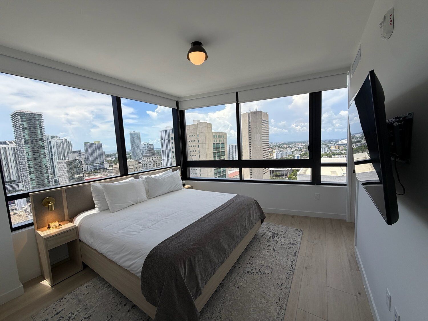 A bedroom with a large bed, a nightstand, a TV mounted on the wall, and a city view through large windows.