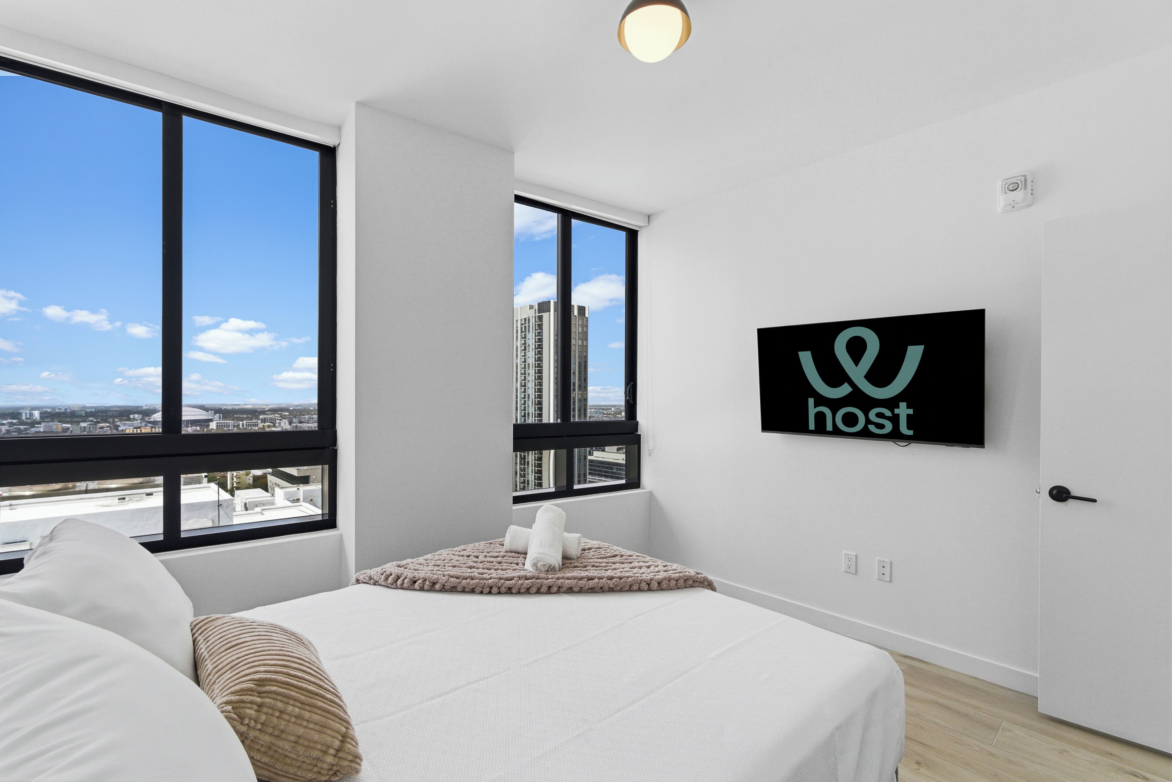 Quarto de hotel moderno com uma cama grande, vista da cidade através de uma janela e uma TV na parede exibindo a palavra 'host'.