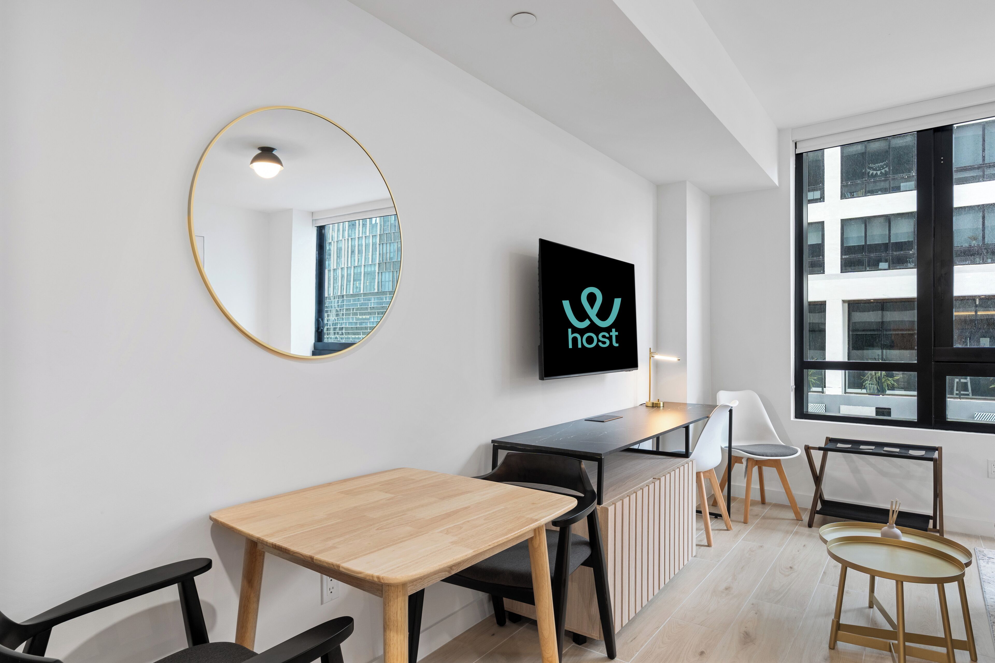 Una habitación moderna y bien iluminada con una mesa de madera, sillas negras, un espejo redondo y un televisor que muestra la palabra "host".