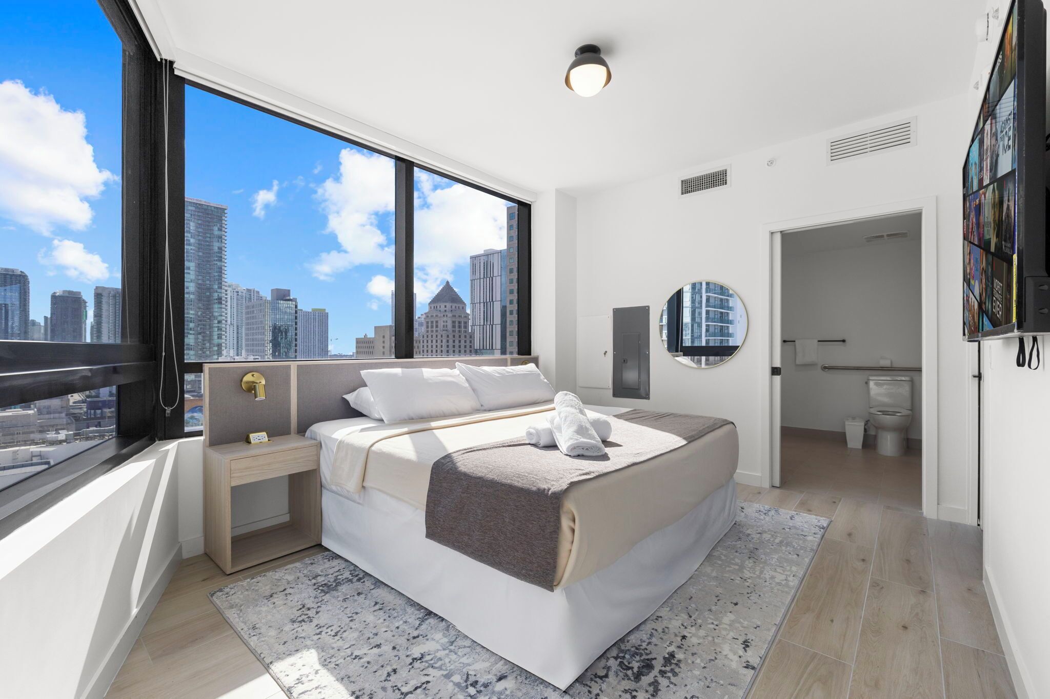 Un dormitorio moderno con una cama grande, una mesita de noche, un tocador, un espejo y vistas de la silueta urbana a través de amplias ventanas.