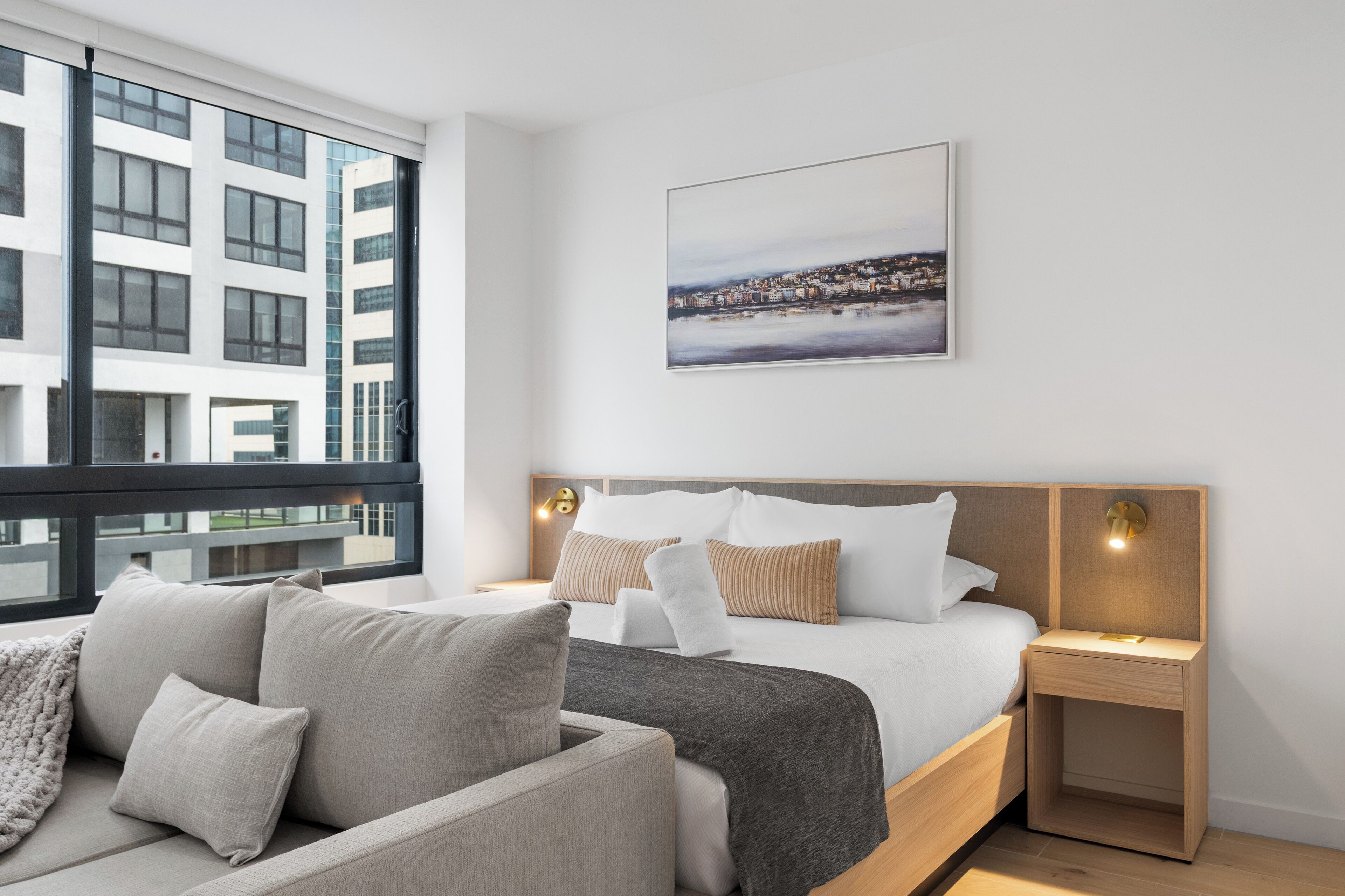 Une chambre d’hôtel moderne avec un grand lit, une table de chevet avec une lampe, une vue sur le paysage urbain à travers de grandes fenêtres et un tableau encadré au mur.