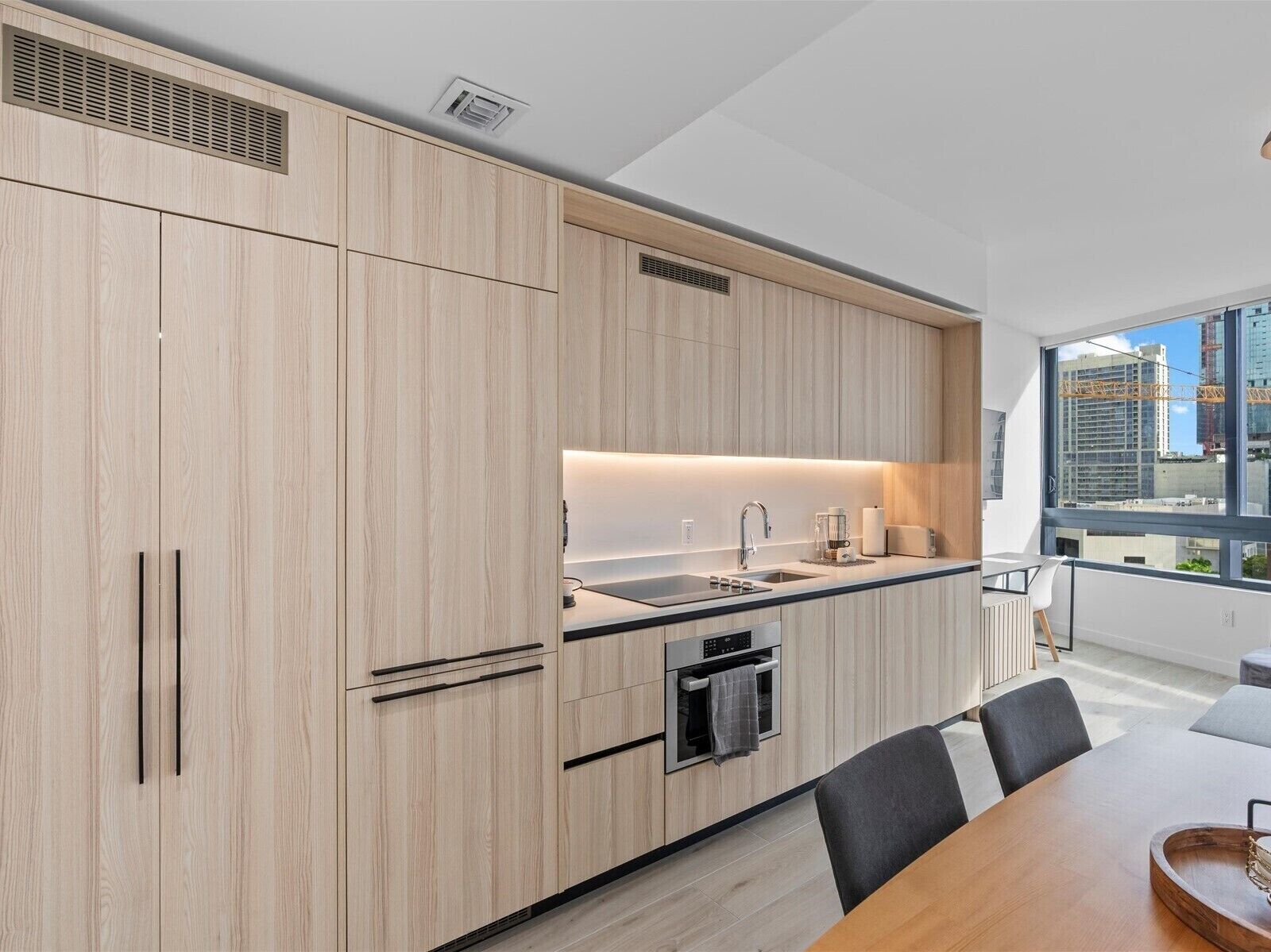 A modern kitchen with light wood cabinetry, a built-in oven, and a large window offering a view of the cityscape.