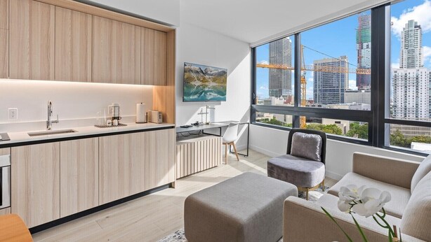 A modern kitchen with a large island, a dining area with a sofa and ottoman, and a large window with a city view. - Downtown Building - Scenic waterfront walking (Miami)