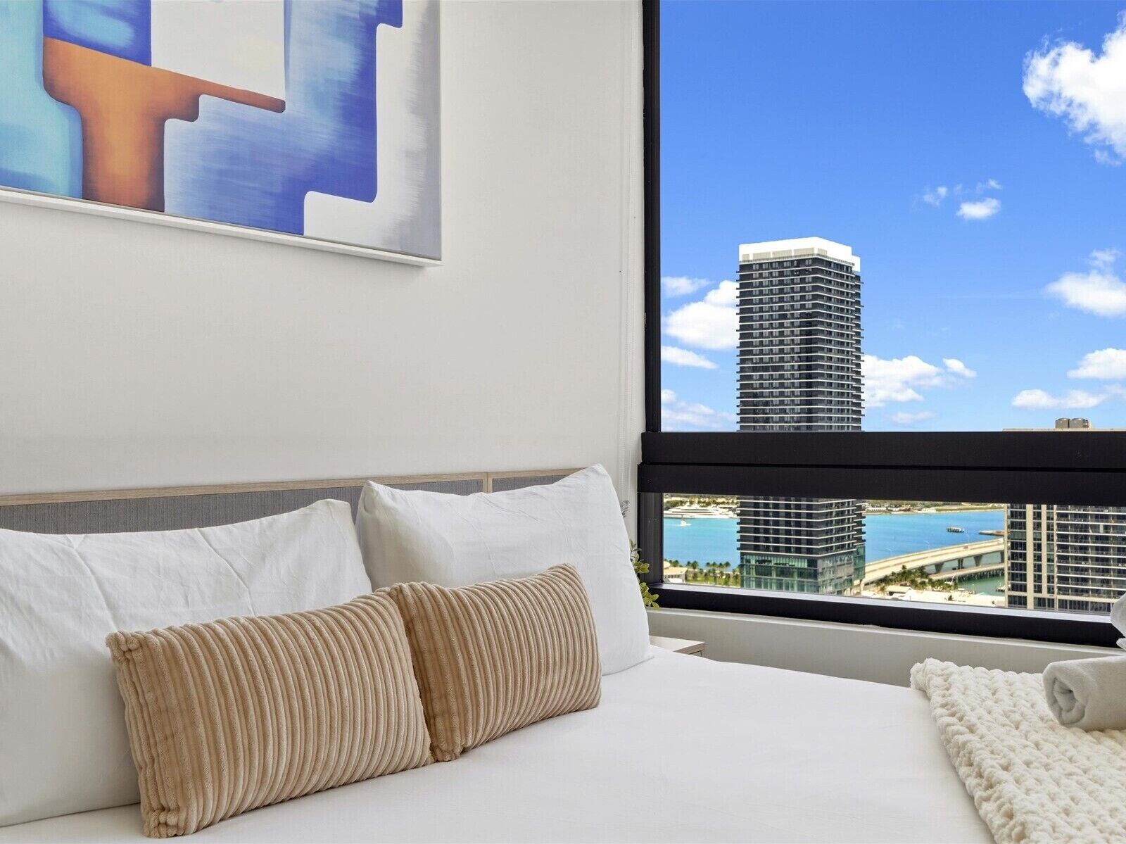 Une chambre d’hôtel avec une grande fenêtre, un lit avec du linge de maison blanc et une vue sur un immeuble imposant et sur l’eau.