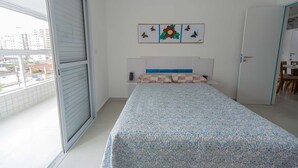 A bedroom with a bed, a window with blinds, a door, and a small table with a chair. - Apto com Garagem e Varanda a 450m da Praia (Praia Grande)