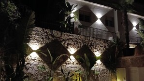Un muro de piedra con plantas e iluminación, un balcón con barandillas y un edificio al fondo.