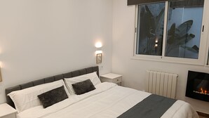 Un dormitorio con una cama, paredes blancas, una mesita de noche, una lámpara de pared, una ventana con vistas a la vegetación y una chimenea.